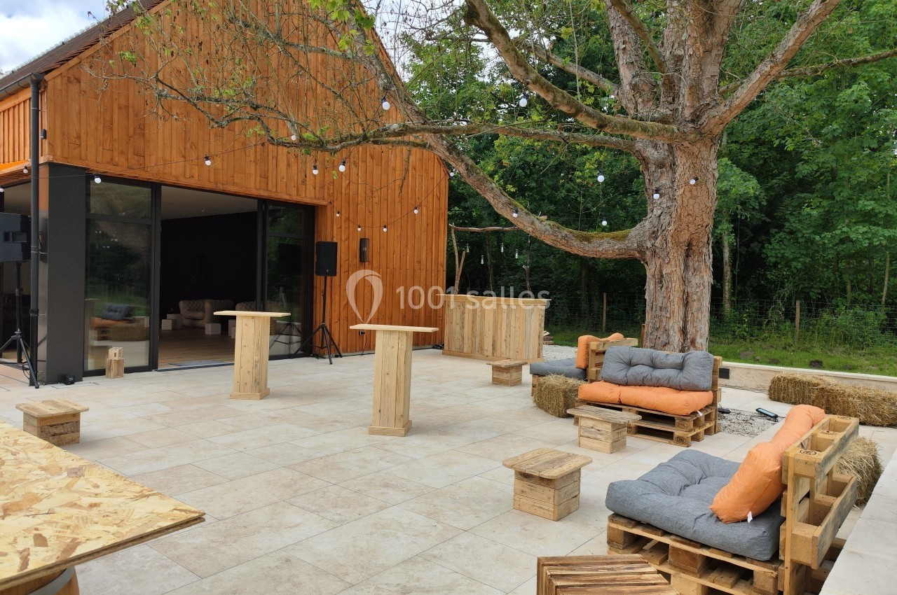 Terrasse aménagée avec mobilier en bois, coussins orange et un grand arbre, devant un bâtiment en bois.