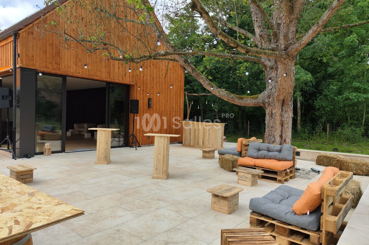 Terrasse aménagée avec mobilier en bois, coussins orange et un grand arbre, devant un bâtiment en bois.