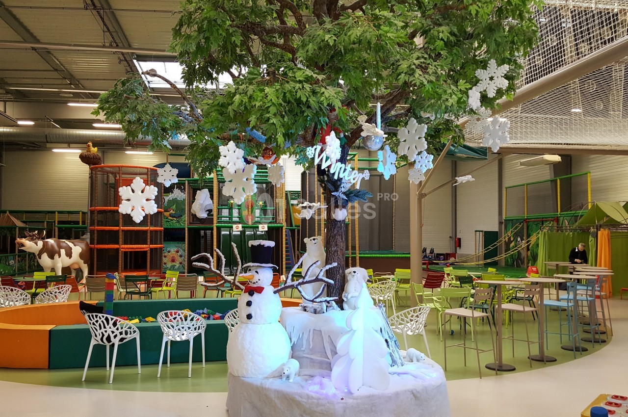 Aire de jeux intérieure avec décorations hivernales, incluant un arbre central, un bonhomme de neige et des tables colorées.