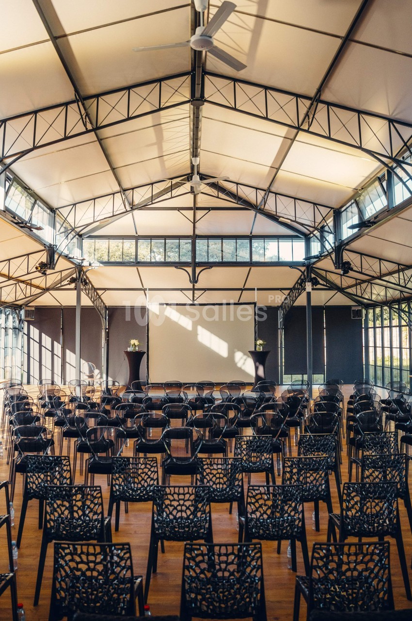 Salle lumineuse avec des rangées de chaises noires devant un écran blanc, sous une structure métallique et un toit en toile.