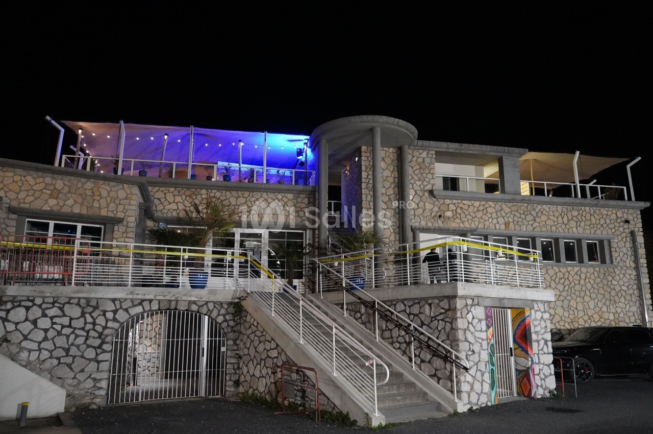 Façade d'un bâtiment en pierre avec terrasses éclairées et escalier central, photographié de nuit.