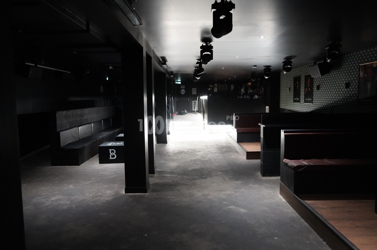 Salle sombre avec banquettes noires et rouges, éclairée par une lumière naturelle provenant d'une porte au fond.