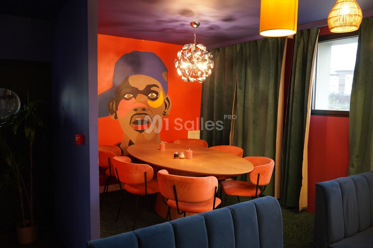 Salle colorée avec une grande table ronde entourée de chaises orange, mur décoré d'un portrait stylisé et luminaires…