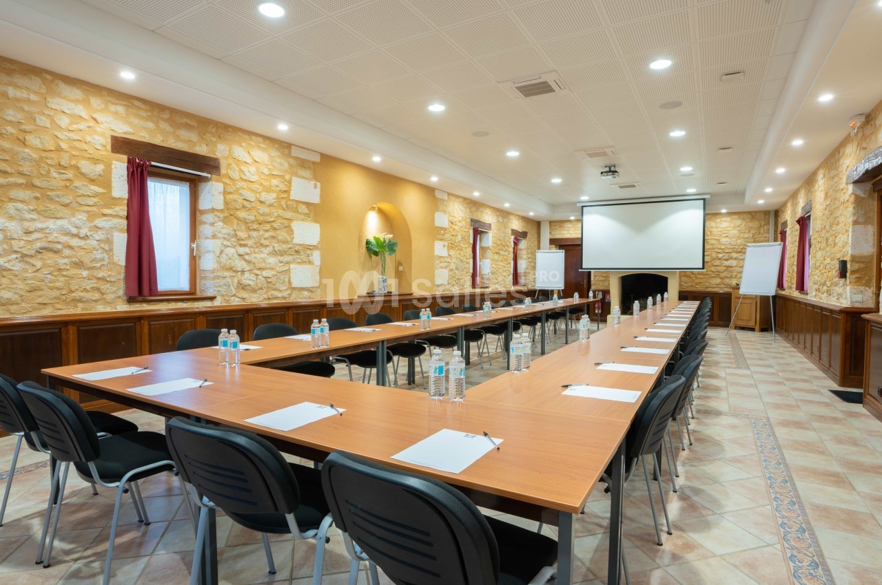 Salle de réunion avec tables disposées en U, chaises, écran de projection, blocs-notes et bouteilles d'eau.