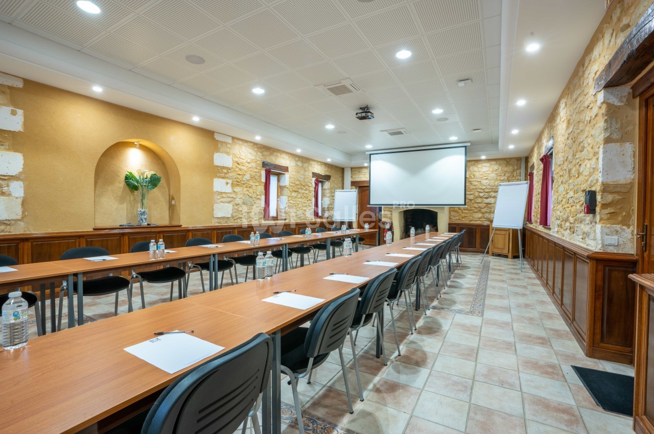 Salle de réunion lumineuse avec tables en U, chaises, écran de projection, paperboards et murs en pierre apparente.