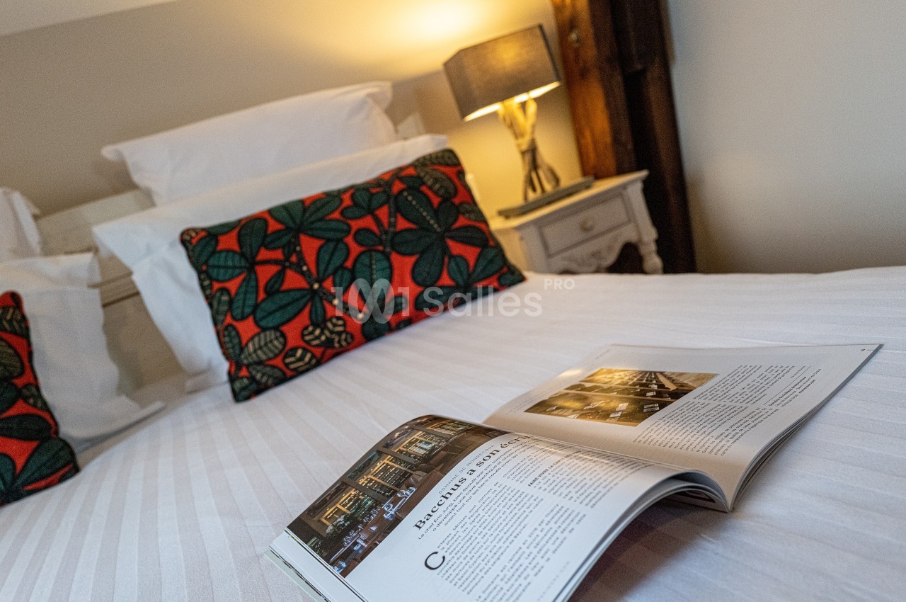 Lit avec draps blancs, coussins à motifs colorés, lampe allumée sur une table de chevet et magazine ouvert au premier plan.