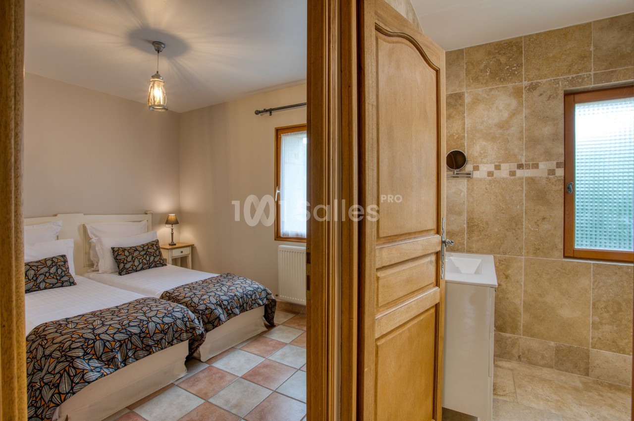 Chambre avec deux lits simples et salle de bain attenante, décorée avec des tons neutres et carrelage beige.