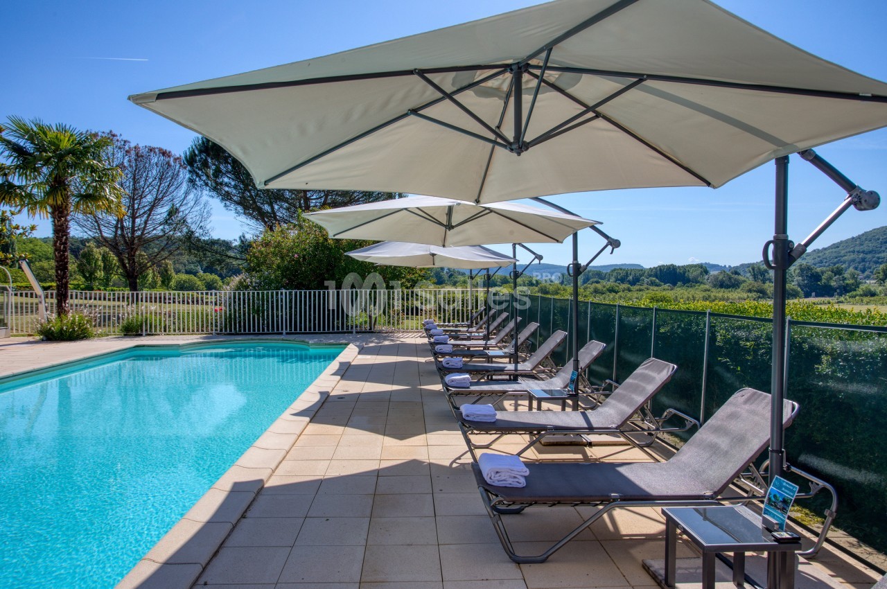 Chaises longues et parasols alignés au bord d'une piscine extérieure entourée de verdure sous un ciel bleu.