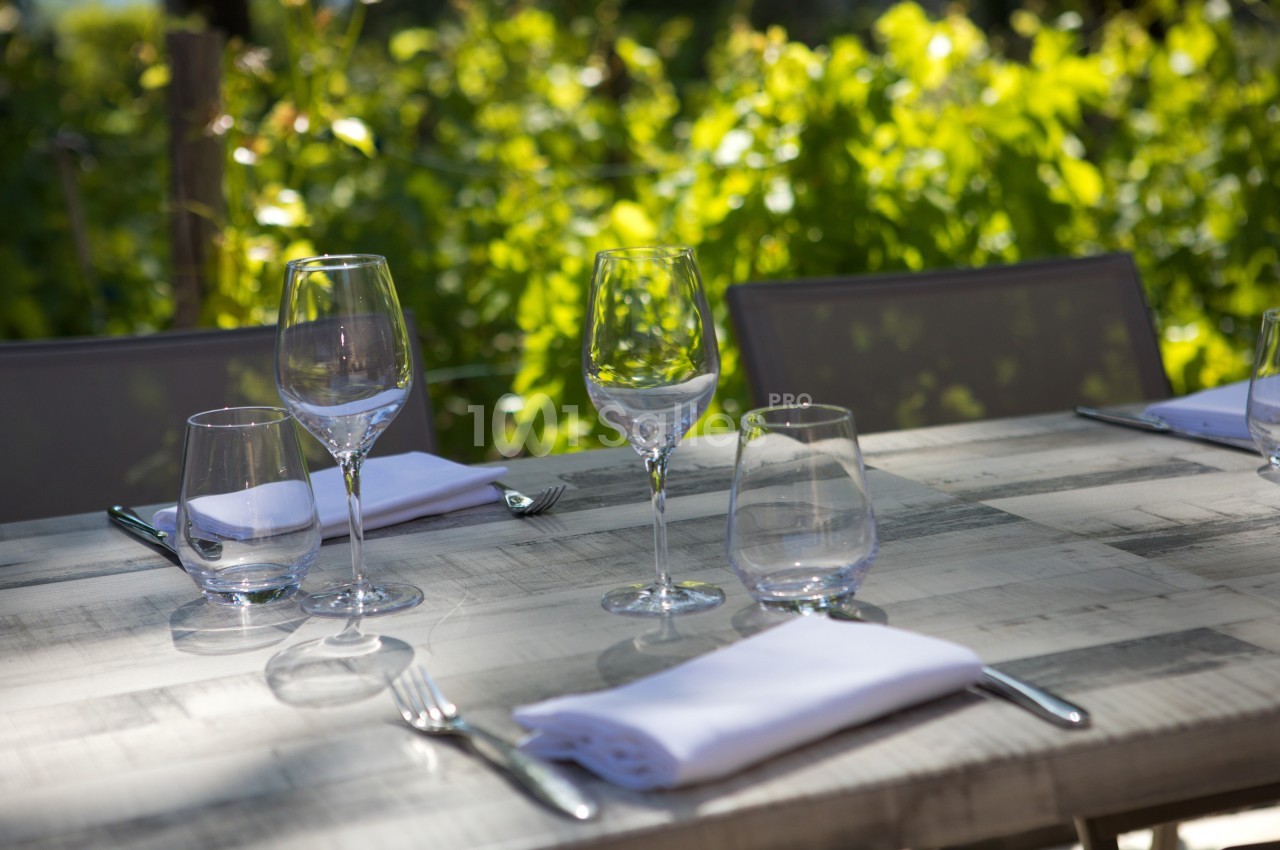 Table en bois dressée avec des verres, des couverts et des serviettes blanches, en extérieur avec un fond de verdure.