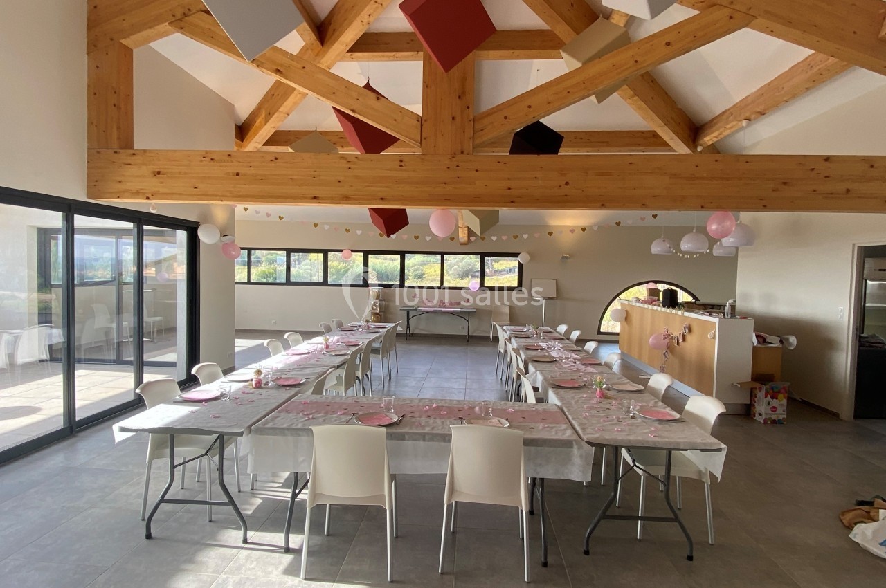 Salle lumineuse décorée pour une réception, avec tables en U dressées et guirlandes suspendues sous une charpente en bois.