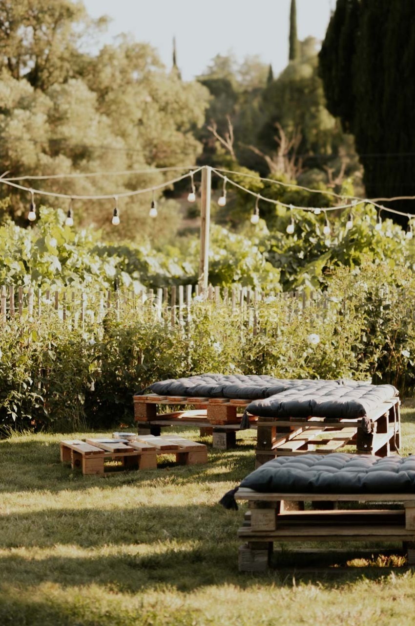 Bancs en palettes avec coussins noirs disposés dans un jardin verdoyant, éclairé par des guirlandes lumineuses.