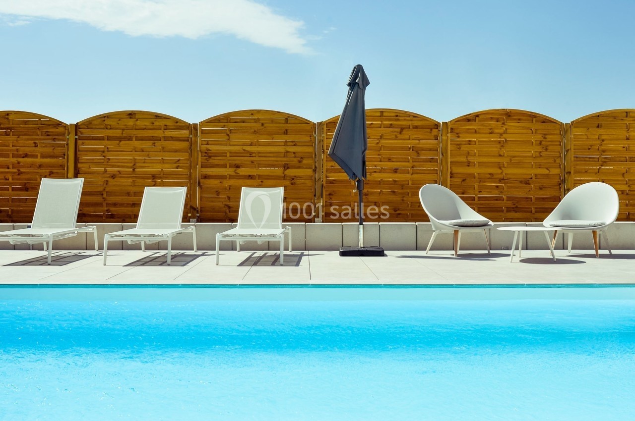 Chaises longues et parasol près d'une piscine, avec une clôture en bois et un ciel dégagé en arrière-plan.