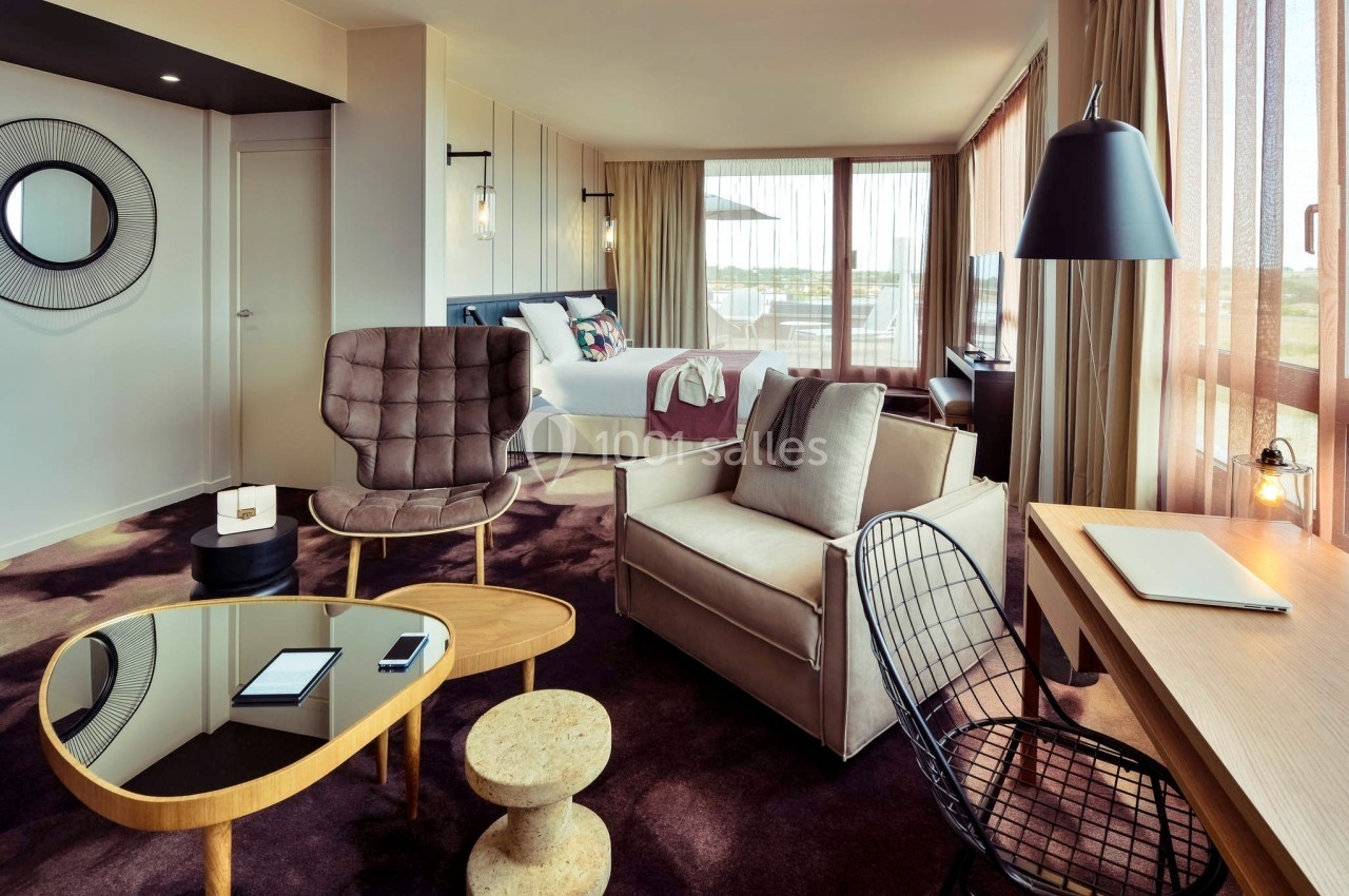 Chambre d'hôtel moderne avec lit, coin salon, bureau, grandes fenêtres et décoration contemporaine.