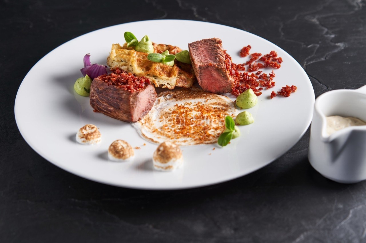 Assiette gastronomique avec viande grillée, purée verte, chips, sauce blanche et décorations raffinées sur une table noire.