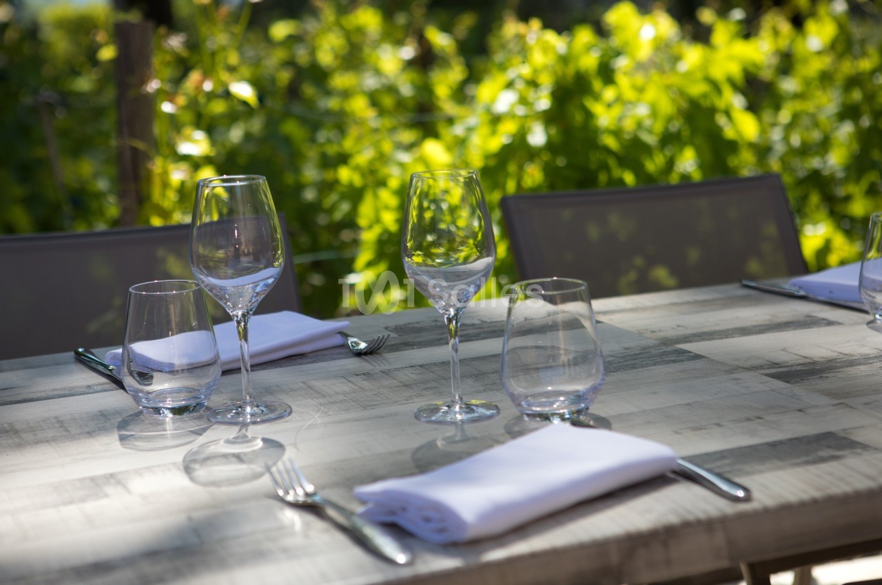 Table en bois dressée avec des verres, des couverts et des serviettes blanches, en extérieur avec un fond de verdure.