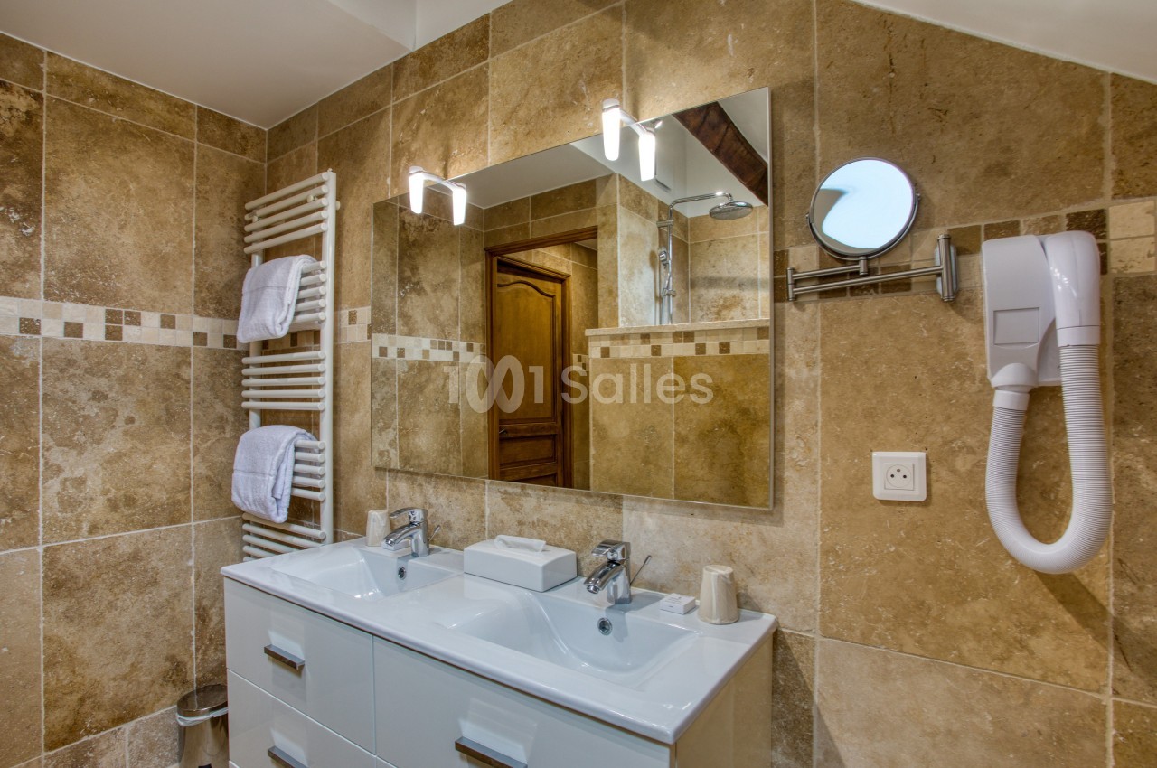 Salle de bain avec double vasque, miroir éclairé, sèche-serviettes, sèche-cheveux et carrelage beige.