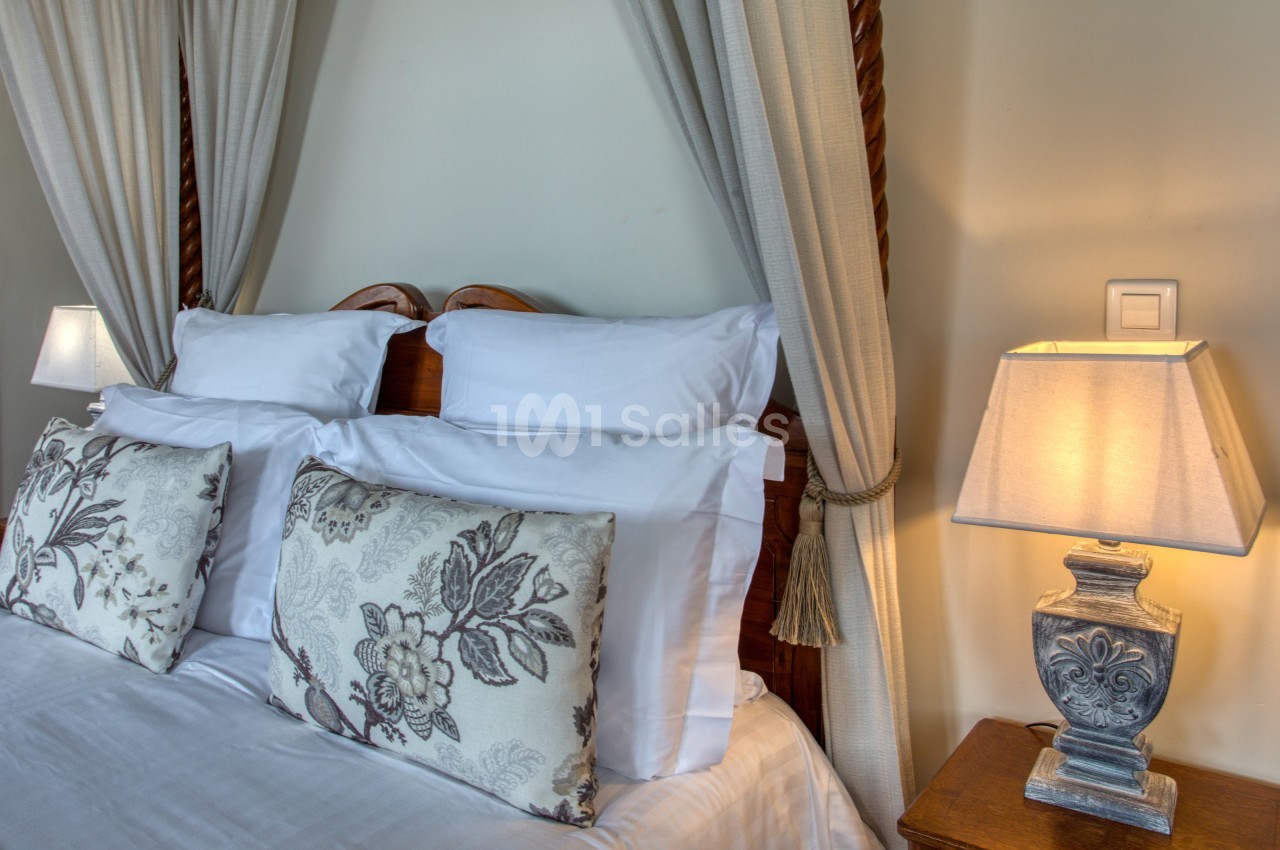 Lit double avec draps blancs, coussins à motifs floraux, lampes de chevet et rideaux décoratifs en arrière-plan.