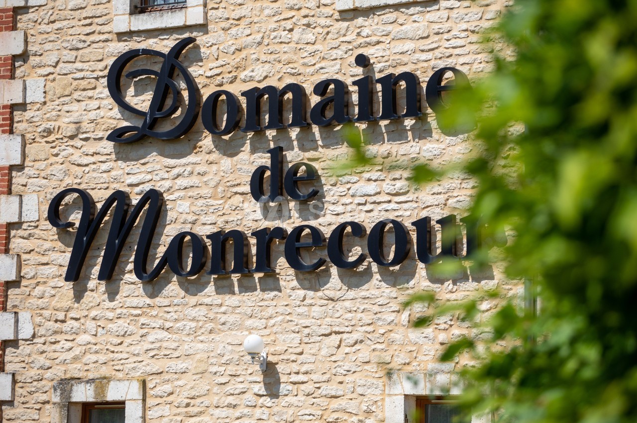 Façade en pierre avec l'inscription ’Domaine de Monrecour’ en lettres noires, partiellement masquée par des feuillages.