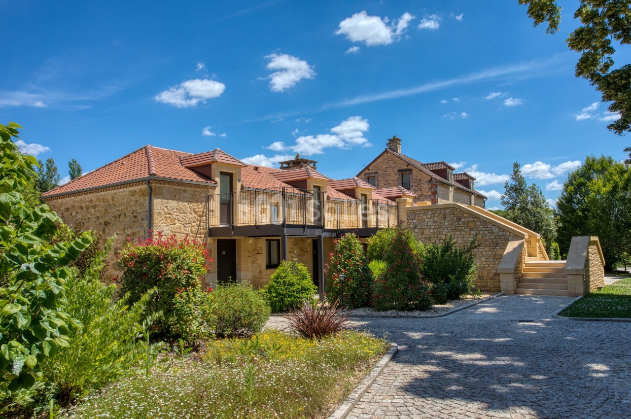 Maison en pierre avec toit en tuiles rouges, entourée de végétation et d'une allée pavée sous un ciel bleu.