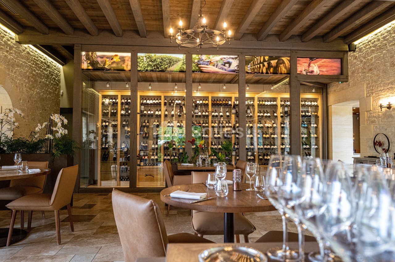 Salle de restaurant élégante avec tables dressées, murs en pierre, plafond en bois et cave à vin vitrée en arrière-plan.