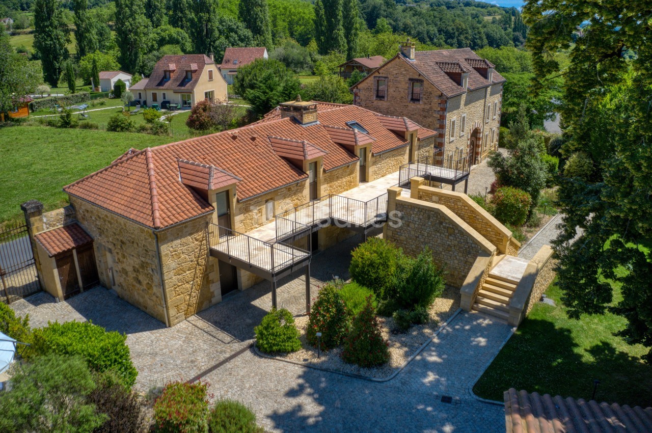 Vue aérienne d'un bâtiment en pierre avec toits en tuiles, entouré de verdure et d'autres habitations.