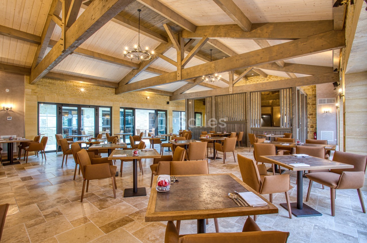 Salle de restaurant lumineuse avec poutres apparentes, tables dressées et chaises en cuir beige.