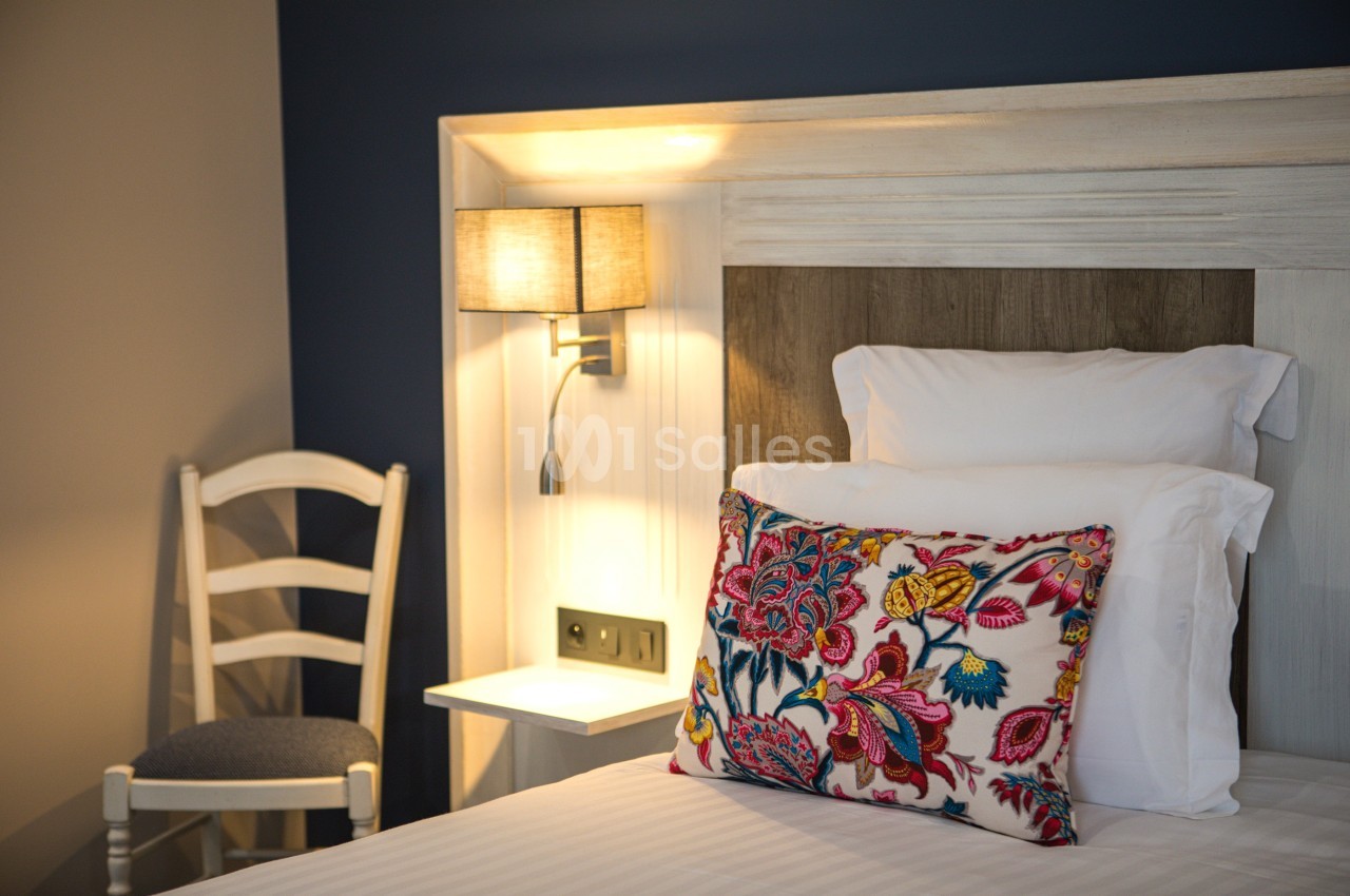 Lit avec une tête de lit en bois, coussin coloré, lampe murale allumée et chaise en bois dans un coin.