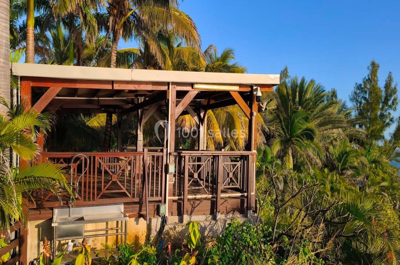 Terrasse en bois entourée de végétation tropicale avec vue partielle sur la mer et ciel dégagé.