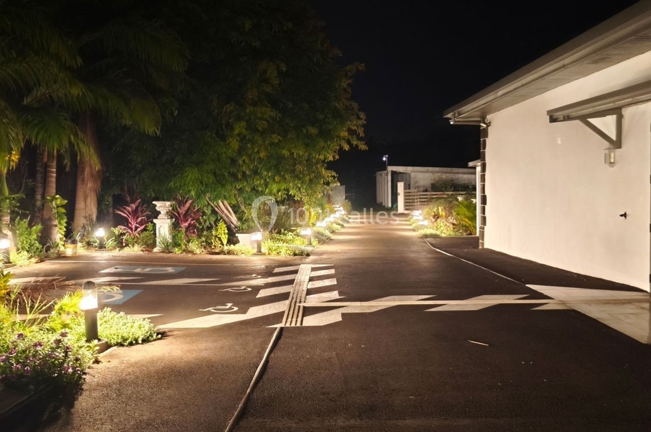 Allée extérieure éclairée la nuit, bordée de végétation et d'un bâtiment blanc avec des marquages au sol.
