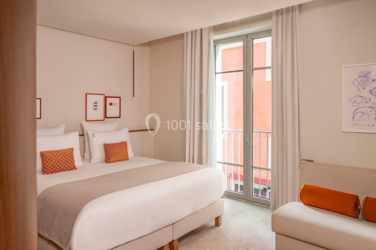 Chambre lumineuse avec un grand lit, canapé, décor minimaliste et fenêtre donnant sur un bâtiment rouge.