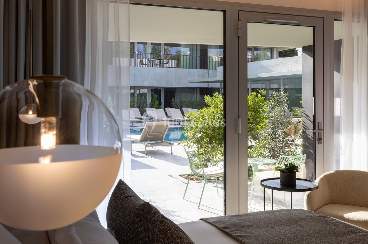 Vue depuis une chambre lumineuse sur une terrasse avec piscine, transats et végétation extérieure.
