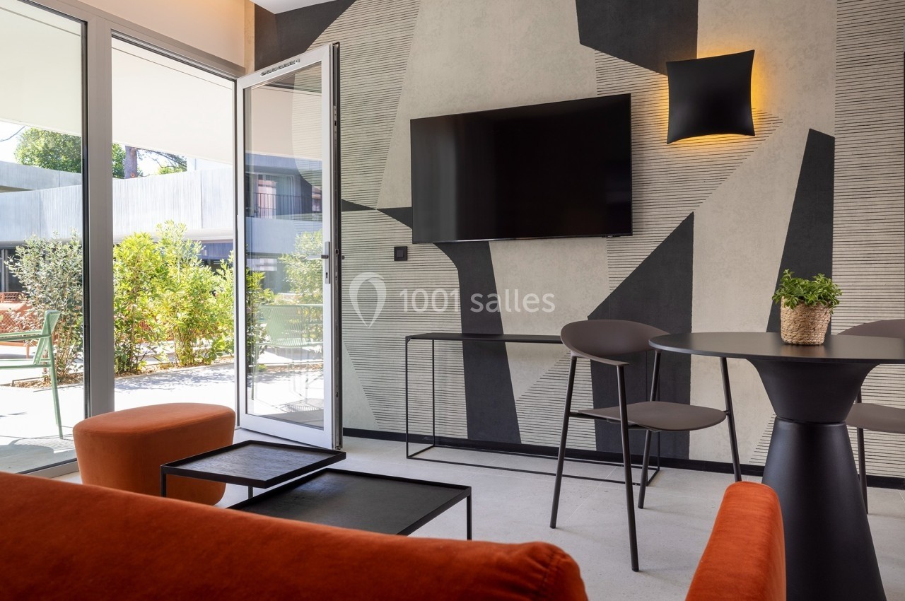 Salon moderne avec canapé orange, table noire, télévision murale et vue sur un jardin par une porte-fenêtre ouverte.