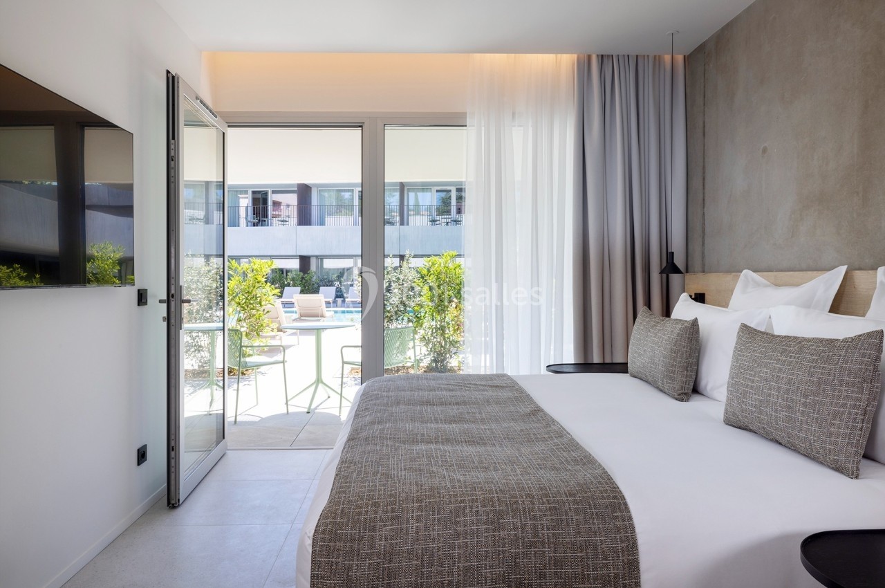 Chambre lumineuse avec lit double, grandes baies vitrées donnant sur une terrasse et vue sur une piscine extérieure.