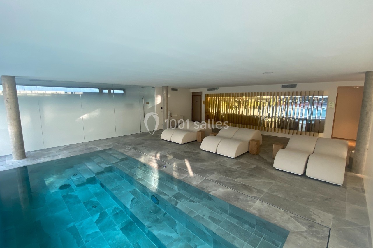 Piscine intérieure avec eau turquoise, entourée de chaises longues modernes sur un sol en pierre grise.