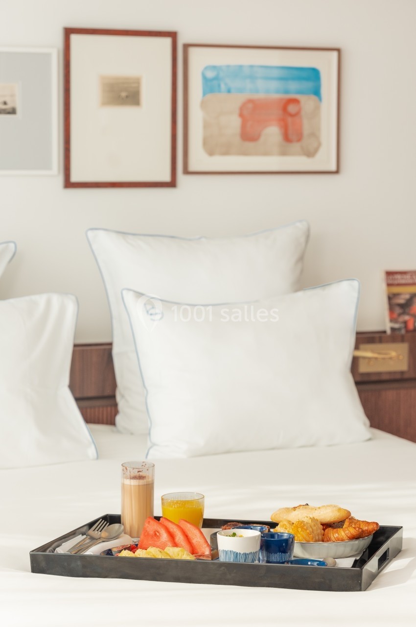 Petit-déjeuner servi sur un plateau avec fruits, viennoiseries et boissons, posé sur un lit aux oreillers blancs.