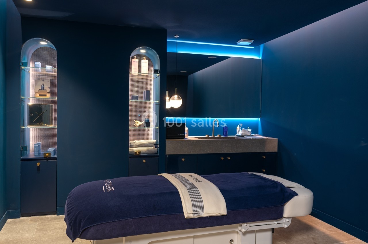 Salle de soins avec table de massage, éclairage tamisé et étagères contenant des produits de beauté.