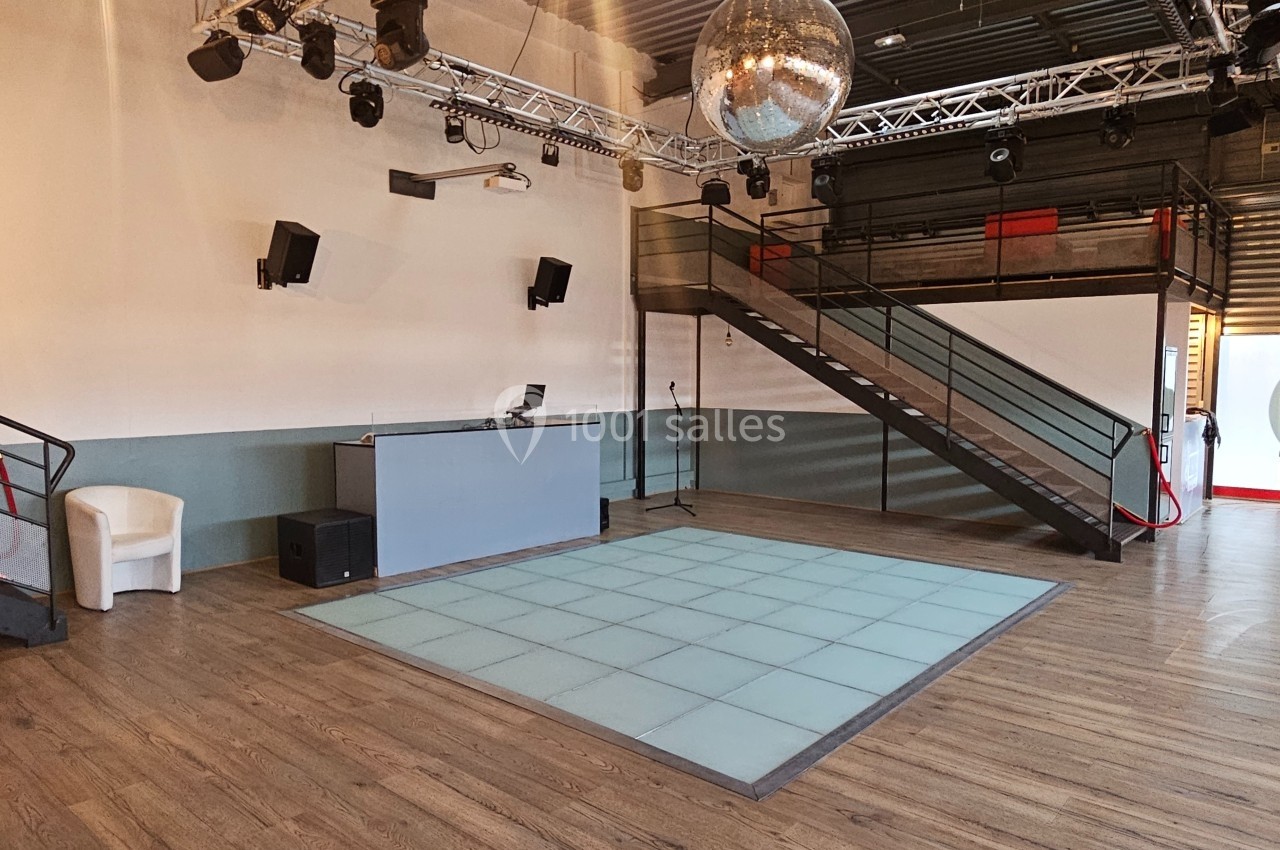 Salle avec piste de danse éclairée, cabine DJ, escalier métallique et boule à facettes suspendue au plafond.