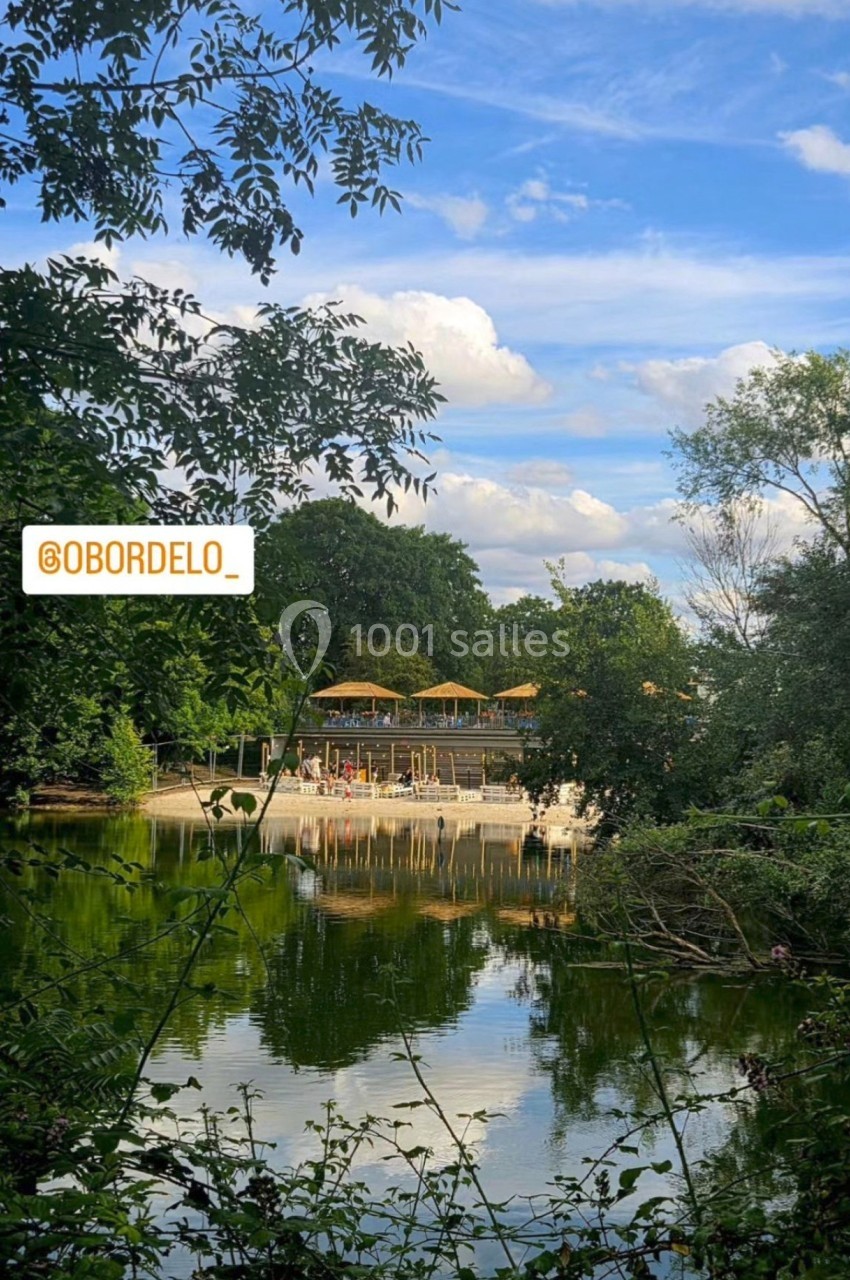 Vue d'un étang entouré de verdure avec une plage aménagée et des parasols en arrière-plan sous un ciel bleu.