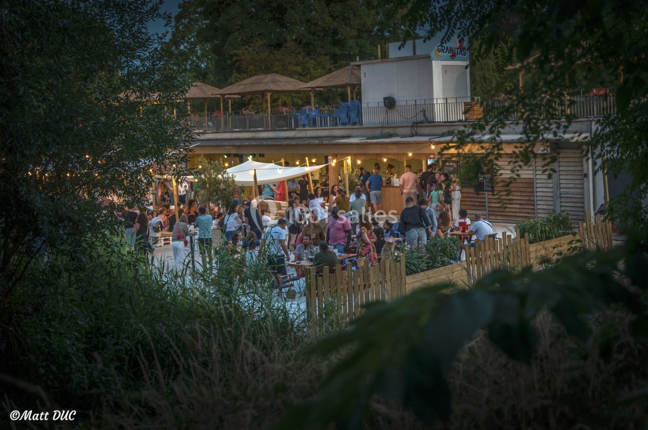 Terrasse animée en soirée avec des tables occupées, éclairages chaleureux et végétation en premier plan.