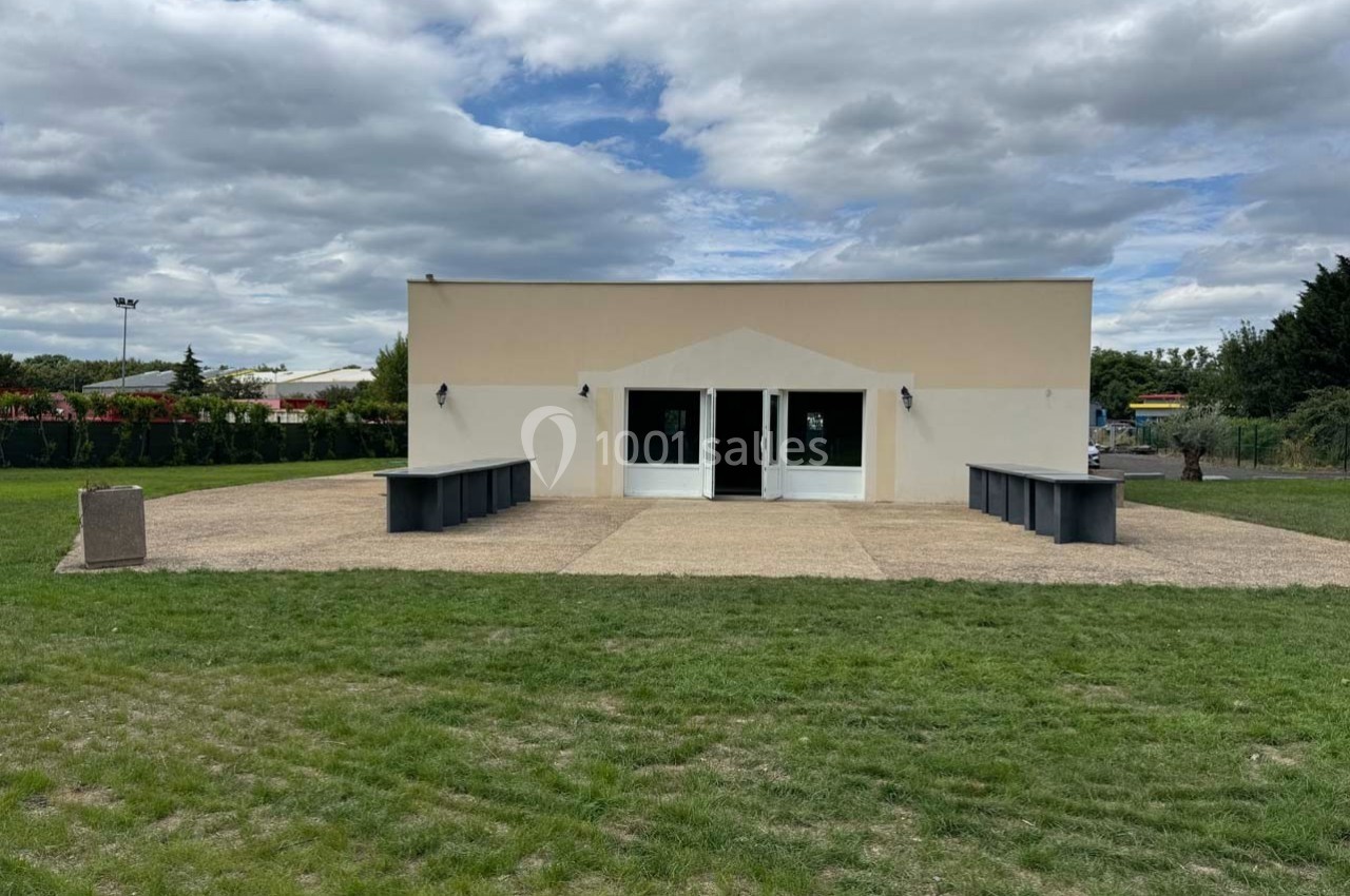 Bâtiment rectangulaire clair avec deux portes vitrées, entouré d'une terrasse pavée et d'une pelouse verte.