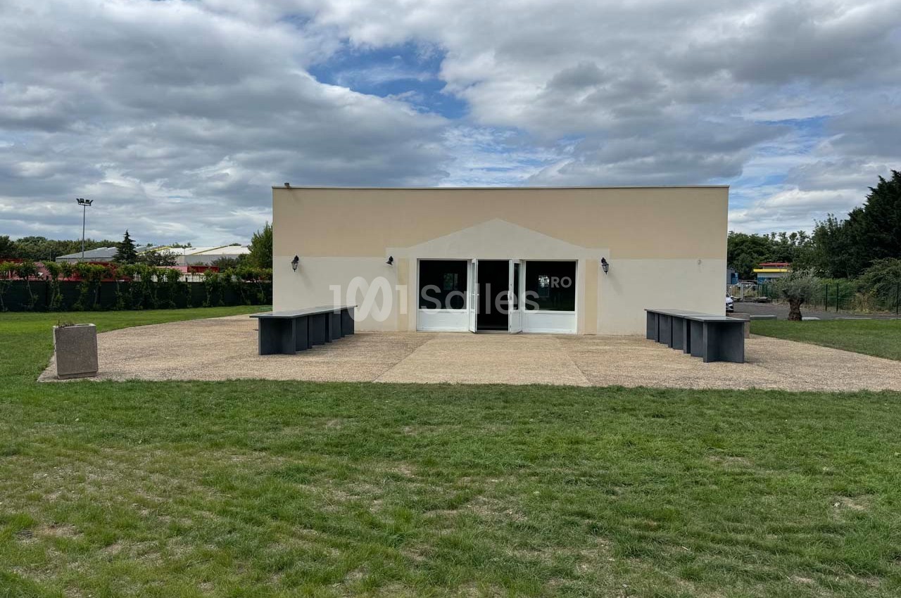 Bâtiment rectangulaire clair avec deux portes vitrées, entouré d'une terrasse pavée et d'une pelouse verte.