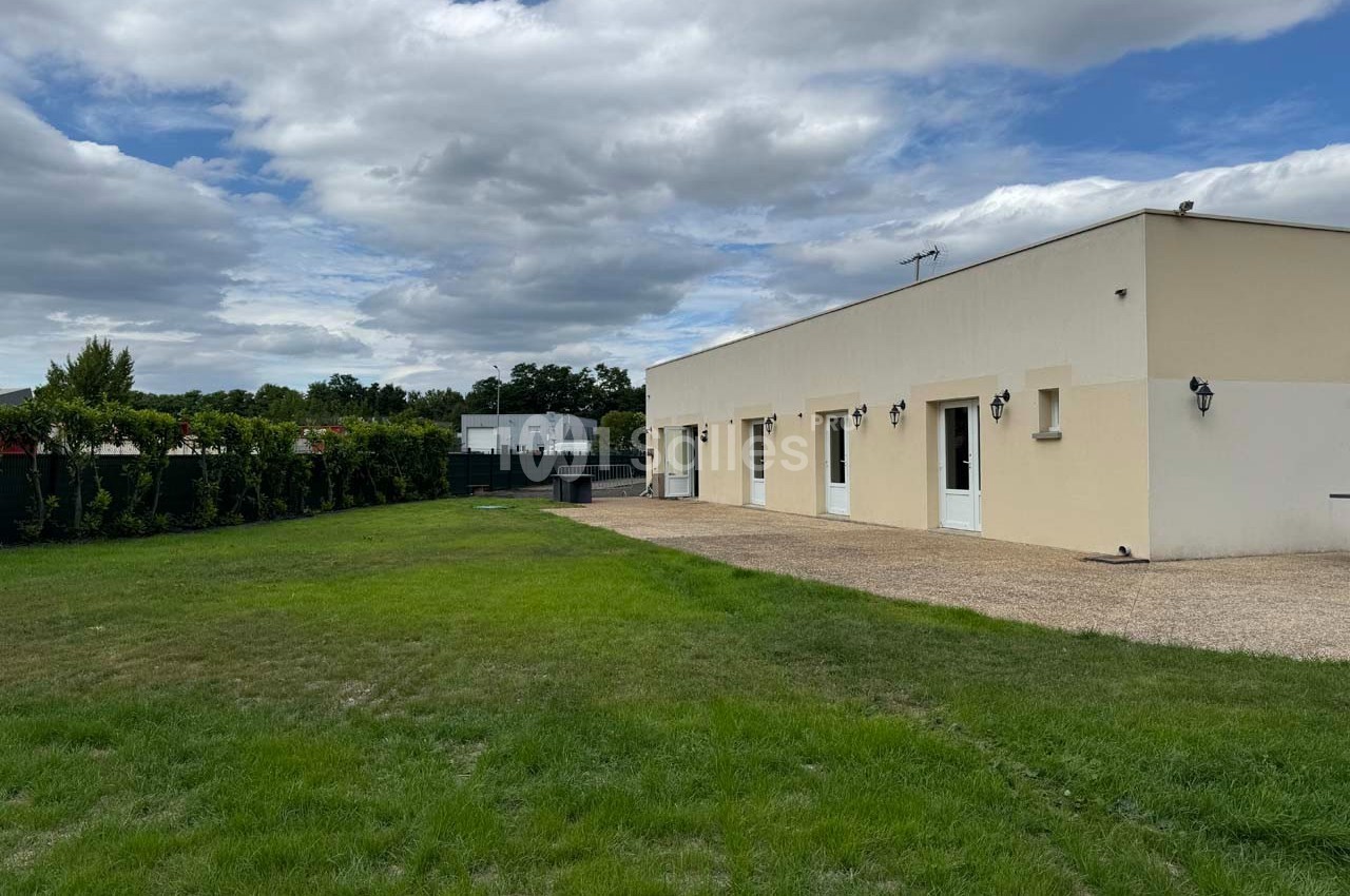 Maison moderne de plain-pied avec façade beige, entourée d'une pelouse et d'une haie, sous un ciel partiellement nuageux.