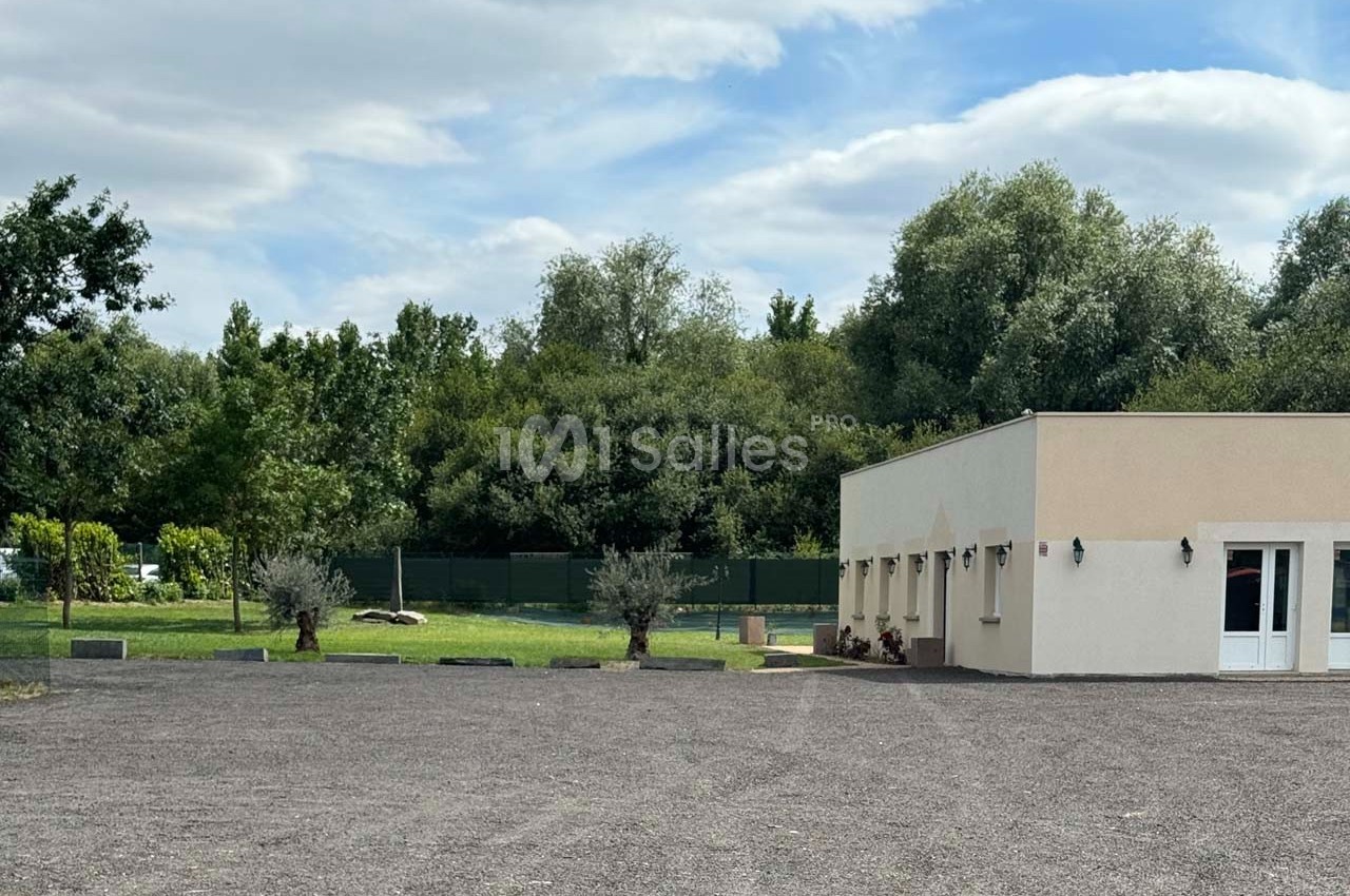 Bâtiment clair avec des murs beige, entouré d'arbres et d'une pelouse, donnant sur un grand parking en gravier.