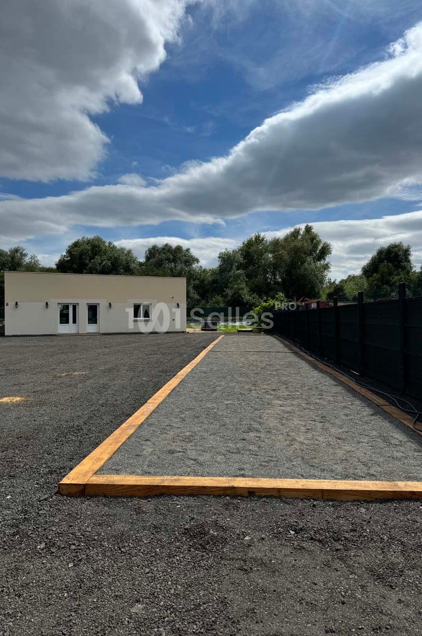 Terrain de pétanque en gravier bordé de bois, situé près d'un bâtiment blanc et entouré de végétation.