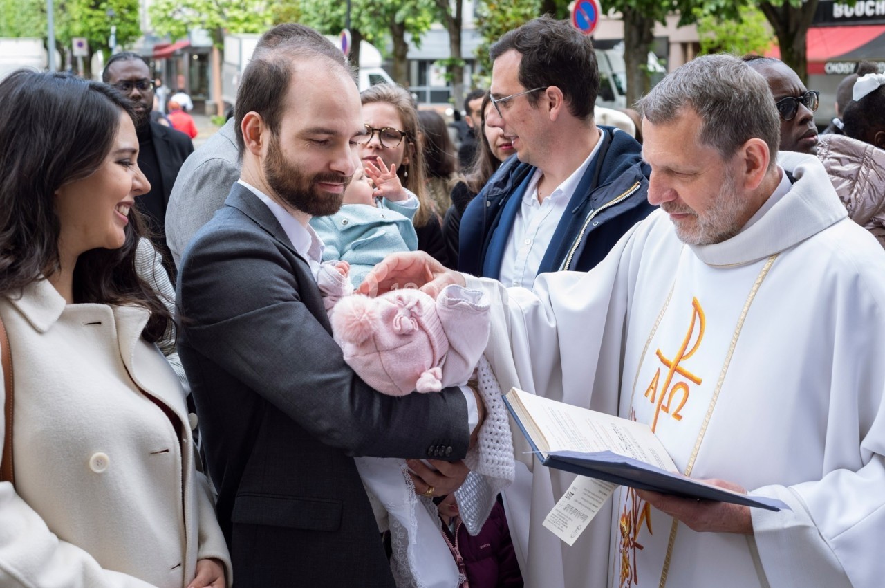 Un prêtre bénit un bébé dans les bras d'un homme, entourés de plusieurs personnes lors d'une cérémonie en extérieur.