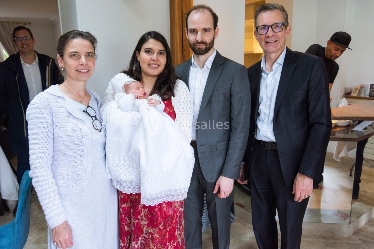 Un groupe de cinq adultes pose ensemble dans une pièce lumineuse, une femme tenant un bébé vêtu de blanc au centre.