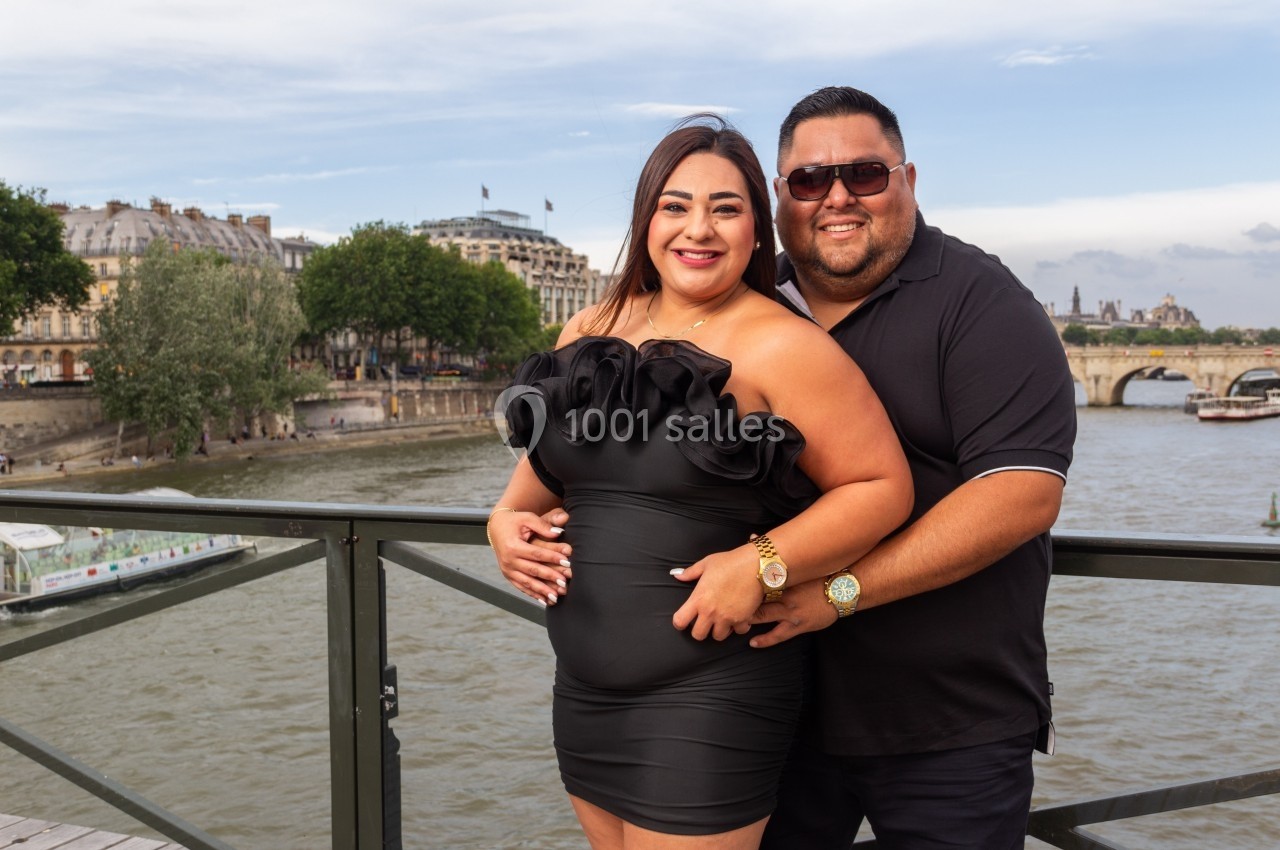 Un couple souriant pose sur un pont avec une rivière et des bâtiments en arrière-plan.