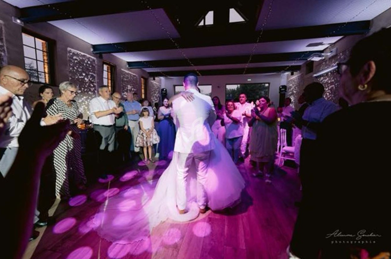 Un couple danse au centre d'une salle éclairée, entouré d'invités applaudissant sur une piste en bois.