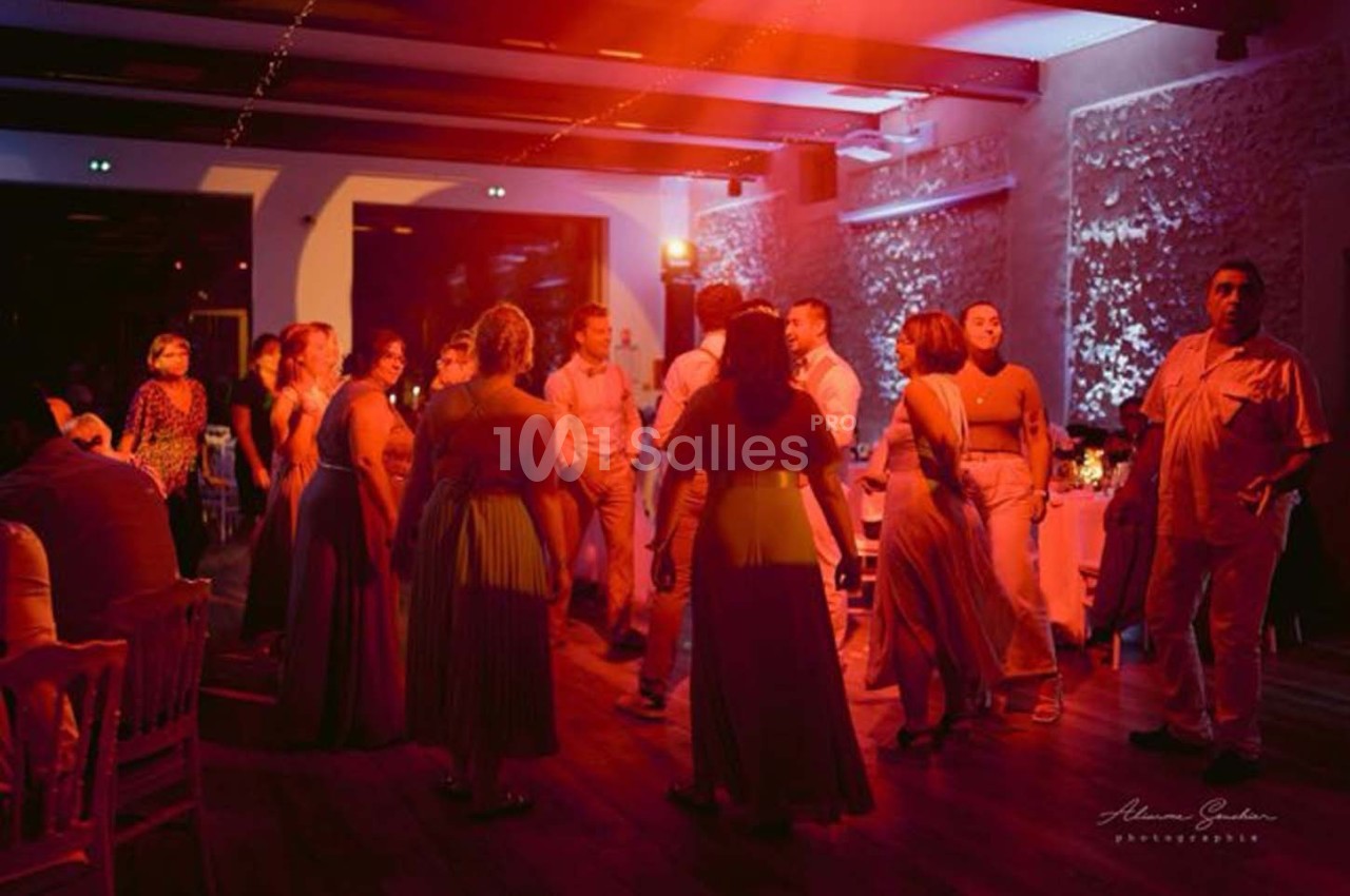 Des personnes dansent dans une salle éclairée par des lumières rouges et blanches, avec des tables en arrière-plan.