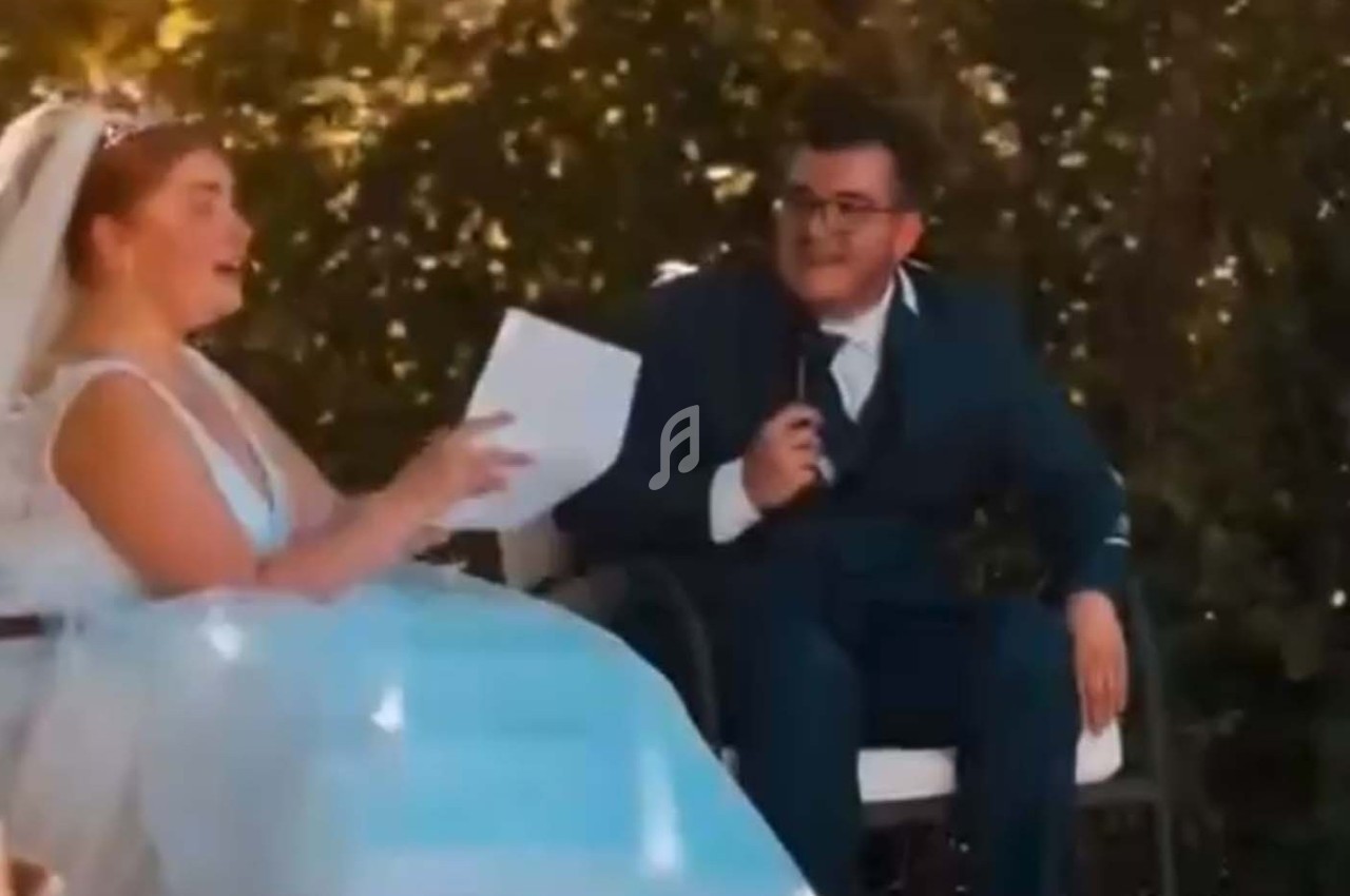 Une mariée en robe blanche et un homme en costume assis échangent lors d'une cérémonie en extérieur.